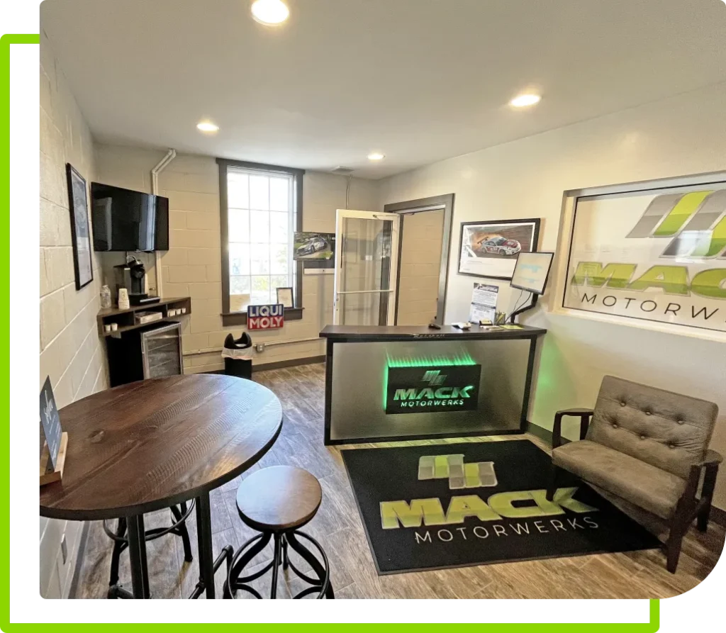 A modern office with a wooden round table and two stools on the left. A reception desk with the logo Mack Motorwerks is on the right. The room has a TV, bright lighting, and large windows in the background.