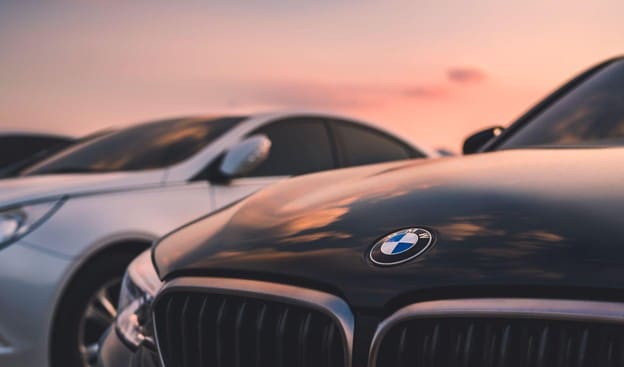 bmw issues, Close-up of a black BMW with its emblem visible, parked alongside other luxury vehicles at sunset.