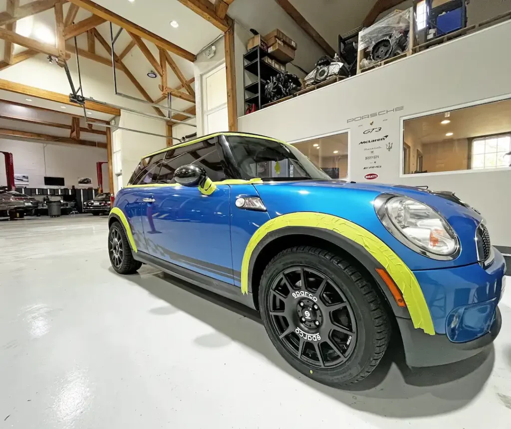 mini cooper repair, auto repair in Jacksonville, FL at Mack Motorwerks. A blue Mini Cooper with protective yellow tape on the edges, parked in a garage with wooden beams and a high ceiling. Various tools and car parts are visible in the background.