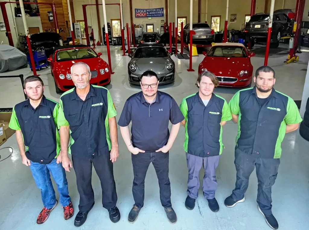 Five men stand in a garage with luxury cars, including a red Bugatti and a black Porsche, in the background. The men wear matching work attire with green and black shirts, and the garage has red lifts and bright lighting.