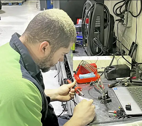 about us, auto repair in Jacksonville, FL at Mack Motorwerks. Man with short hair wearing a green and black shirt, working at a cluttered desk with electronic tools, a laptop, and exposed wires. Hes focused and holding a tool, likely soldering something small and intricate.