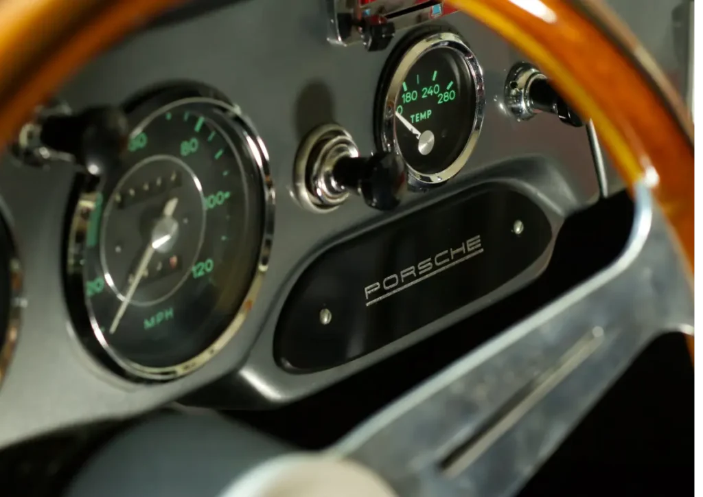 Close-up of a vintage car dashboard featuring a wooden steering wheel and analog gauges. The speedometer shows speeds up to 160 MPH, and a temperature gauge is also visible. The brand name Porsche is displayed prominently on the dashboard.