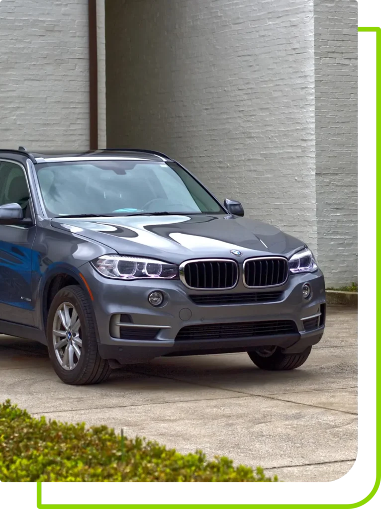 A gray BMW SUV is parked on a concrete driveway in front of a white brick wall. The vehicle is facing slightly to the right, with its headlights illuminated. Green and black borders frame the image. Bushes are visible in the lower left corner.