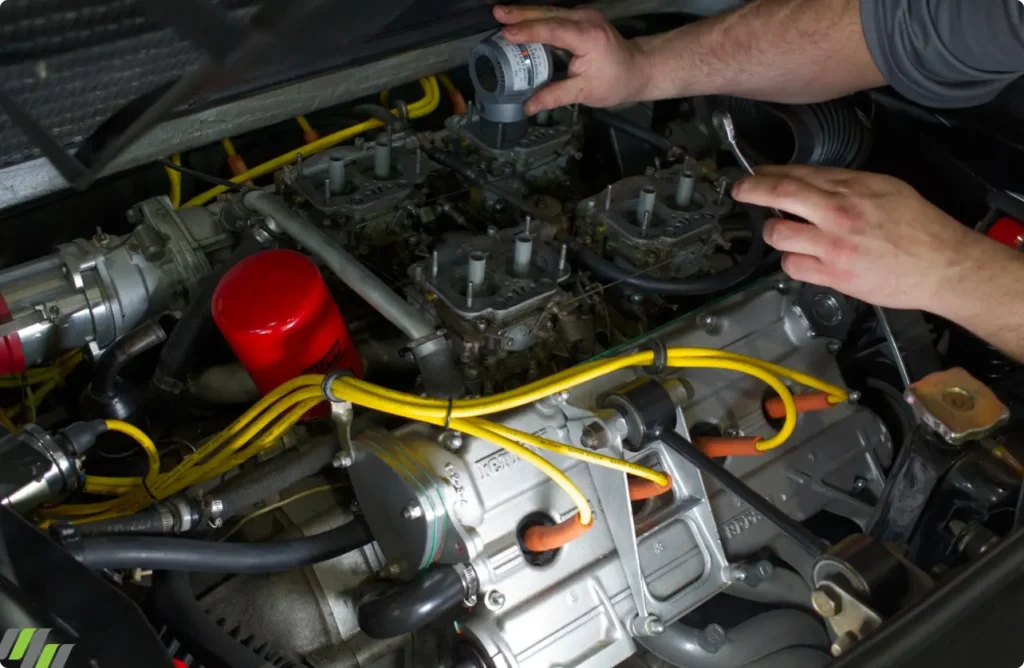 engine diagnostics, auto repair in Jacksonville, FL at Mack Motorwerks. Mechanic working on a car engine, holding a spark plug cap. Yellow wires and various engine components are visible around the mechanics hands, which are reaching into the engine compartment. Tools and parts are scattered around.