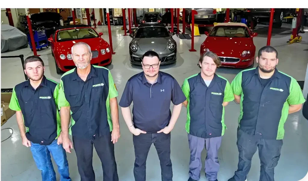 our team, auto repair in Jacksonville, FL at Mack Motorwerks. Five men stand in a garage with luxury cars, including a red Ferrari, silver Porsche, and red Maserati. Three wear matching black and green uniforms, while two others are in casual attire. Red car lifts are visible in the background.