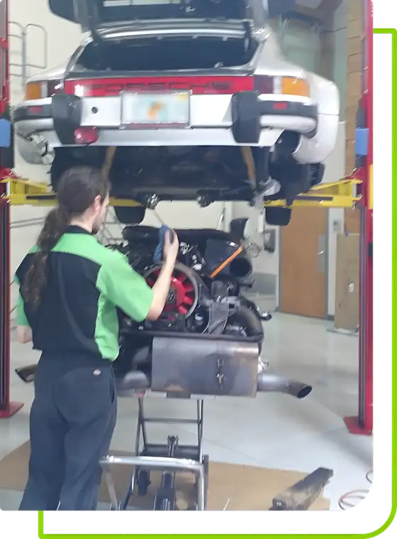 A mechanic with long hair is diligently working on a car engine at Mack Motorwerks, where the vehicle rests on a red hydraulic lift, exposing its undercarriage. Clad in a green and black uniform shirt, the mechanic navigates tools and papers scattered across the floor of this dealership alternative.