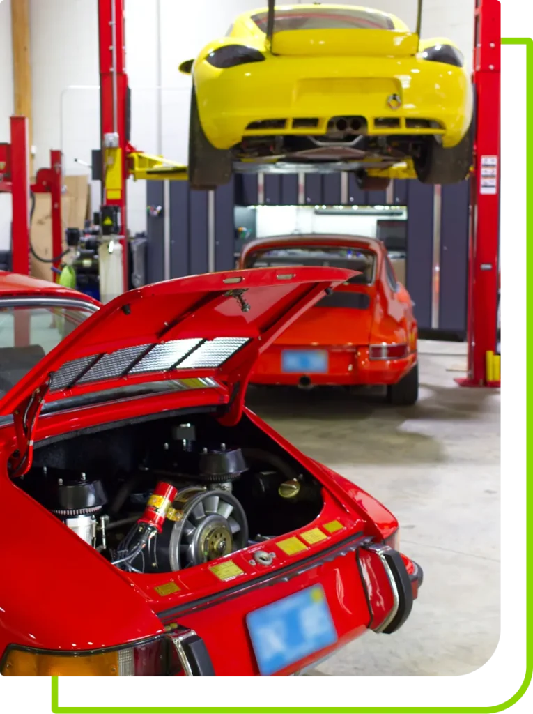 A red vintage sports car with its rear engine compartment open sits in the foreground at Mack Motorwerks, known for expert air-cooled vehicle repair. An orange car is parked nearby, while a yellow one is elevated on a hydraulic lift behind them.
