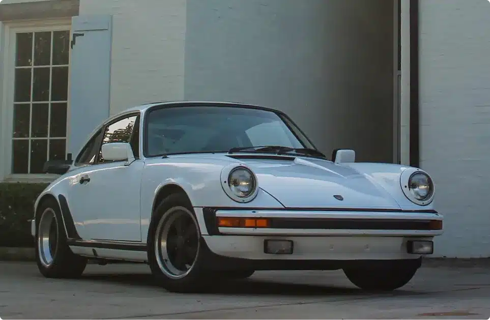 A classic white Porsche 911 sports car is parked in front of a white brick building with a window and an open blue shutter, showcasing the timeless style that Mack Motorwerks brings to every air-cooled vehicle service.
