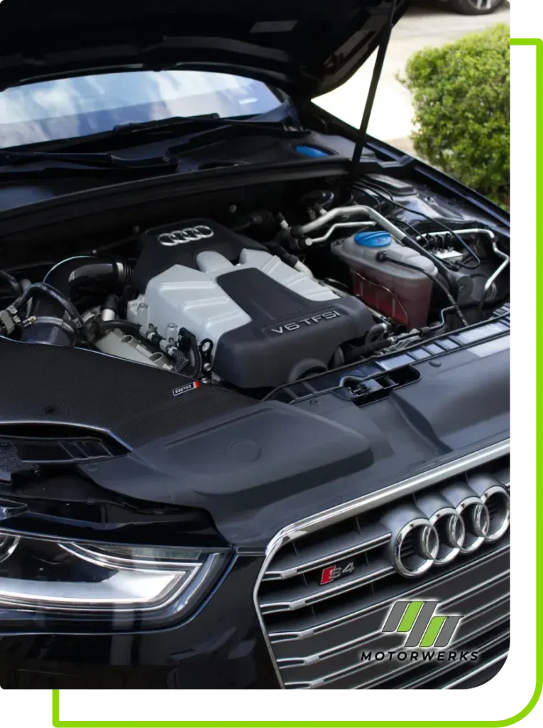 Close-up of the engine bay of an Audi S4, showcasing a V6 TFSI engine with the hood open. The iconic Audi logo gleams on the grille. Against a backdrop of greenery and buildings, this image features Mack Motorwerks branding, highlighting their expertise in European auto repair.