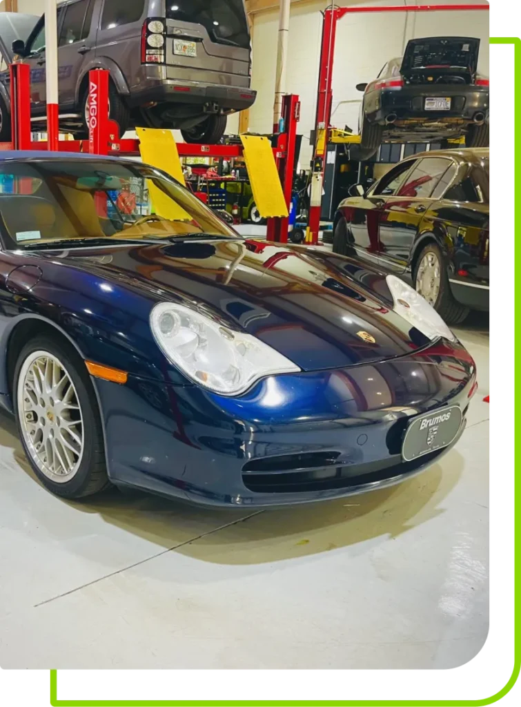 A blue Porsche sports car parked inside an auto repair shop, with other cars lifted on red hydraulic car lifts in the background. Bright lighting reflects off the hood—showcasing this shop as a dealership alternative for auto repair services.