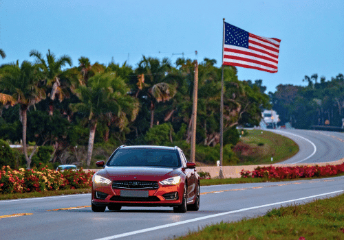 Red car driving on a Florida road with palm trees and a large American flag beside the highway.